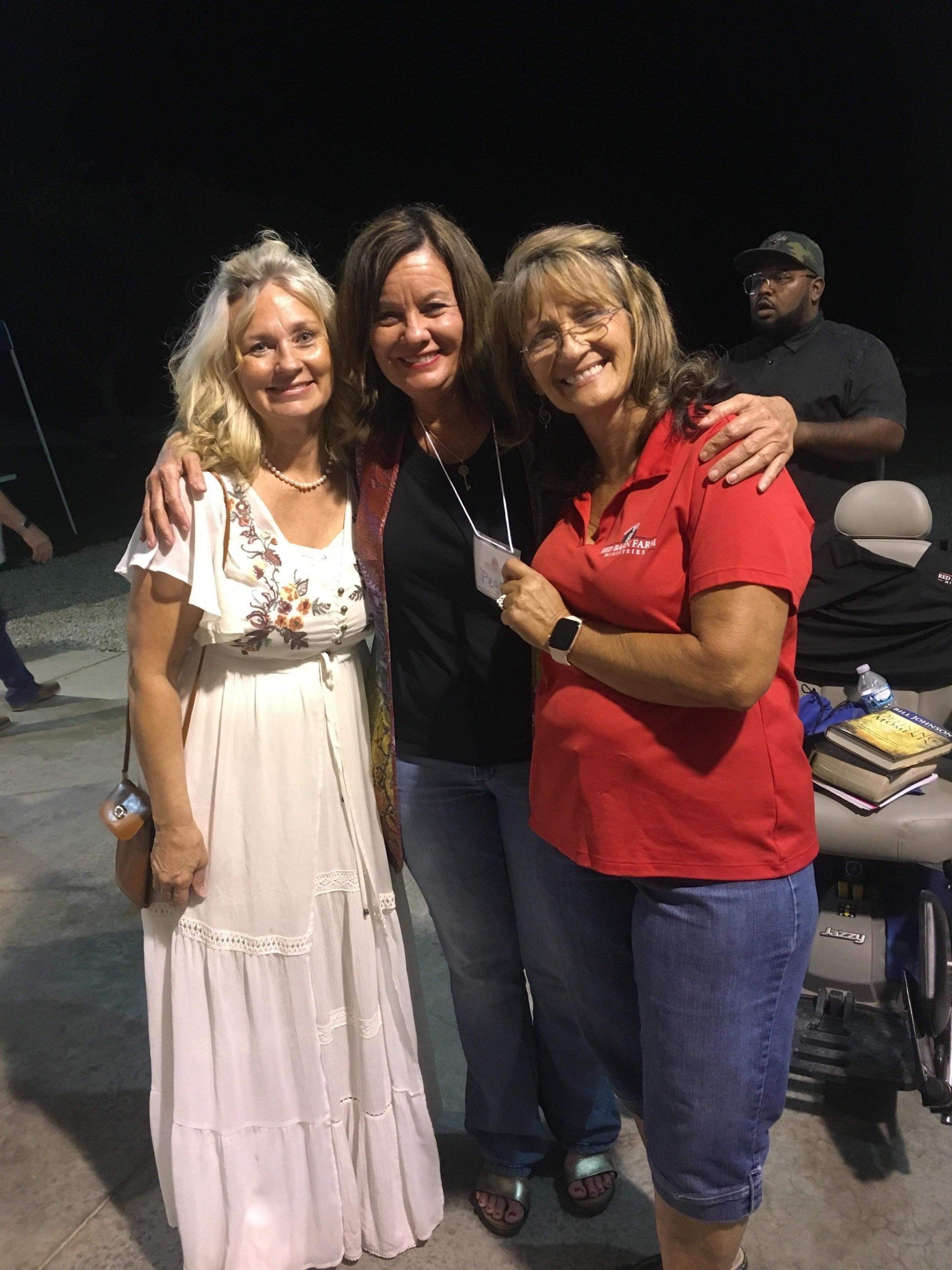 Three women smiling, arms around each other, wearing casual clothes. Outdoor setting, night.