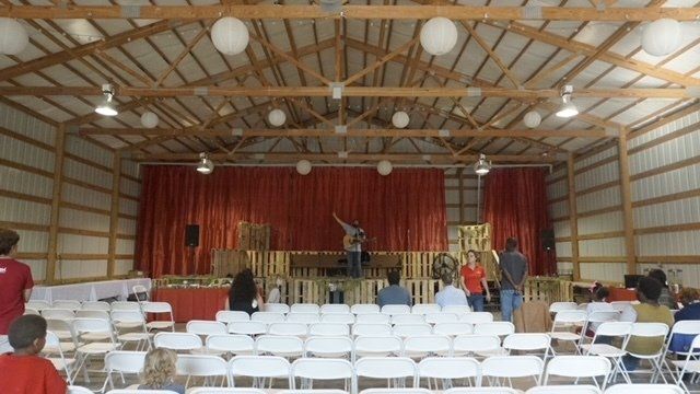 Event in a barn: band on stage with red curtain, people in white chairs, and paper lanterns.