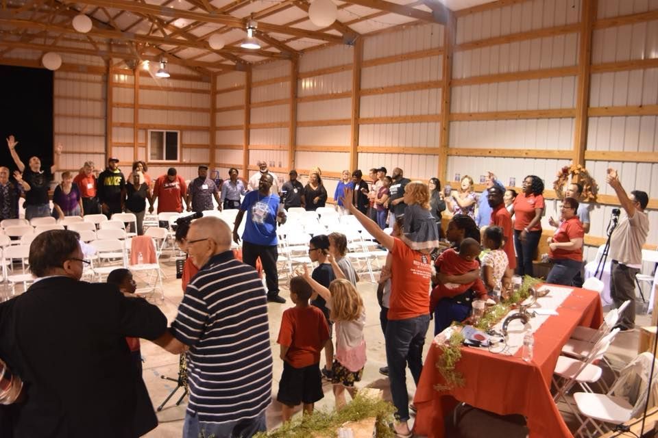 People inside a large building with their hands raised. White chairs and a red-covered table are visible.