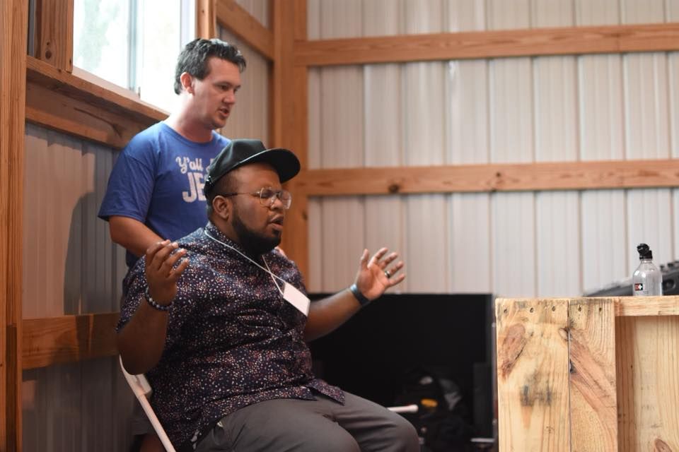 Man seated with arms raised, speaking indoors, another man stands behind him.