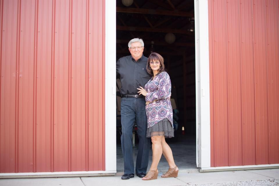 Couple standing in red barn doorway. Woman in dress and boots, man in black shirt and pants.