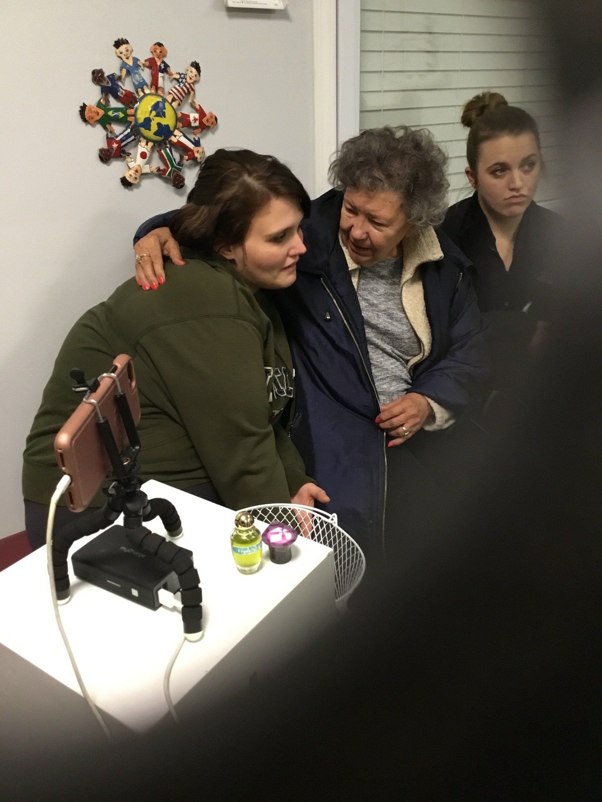 Woman embraces older person; another watches. Smartphone on tripod, objects on table, floral decoration.
