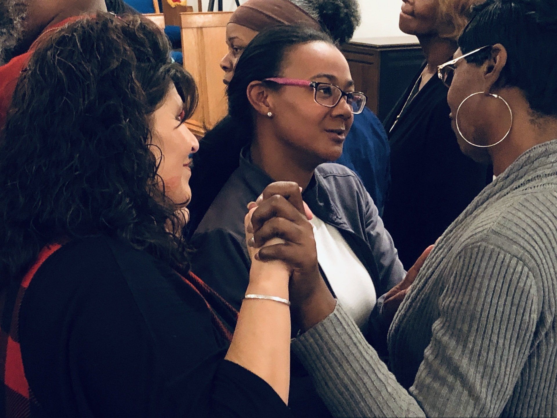 Three women holding hands, talking. One wears glasses, smiles. Indoor setting, soft lighting.