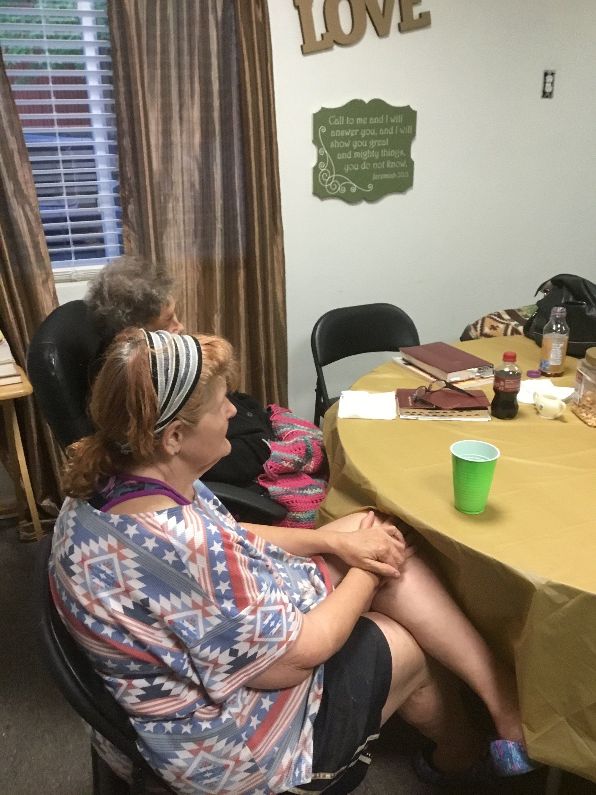 Woman in a colorful top sits at a table with another person. A cup sits on the table.