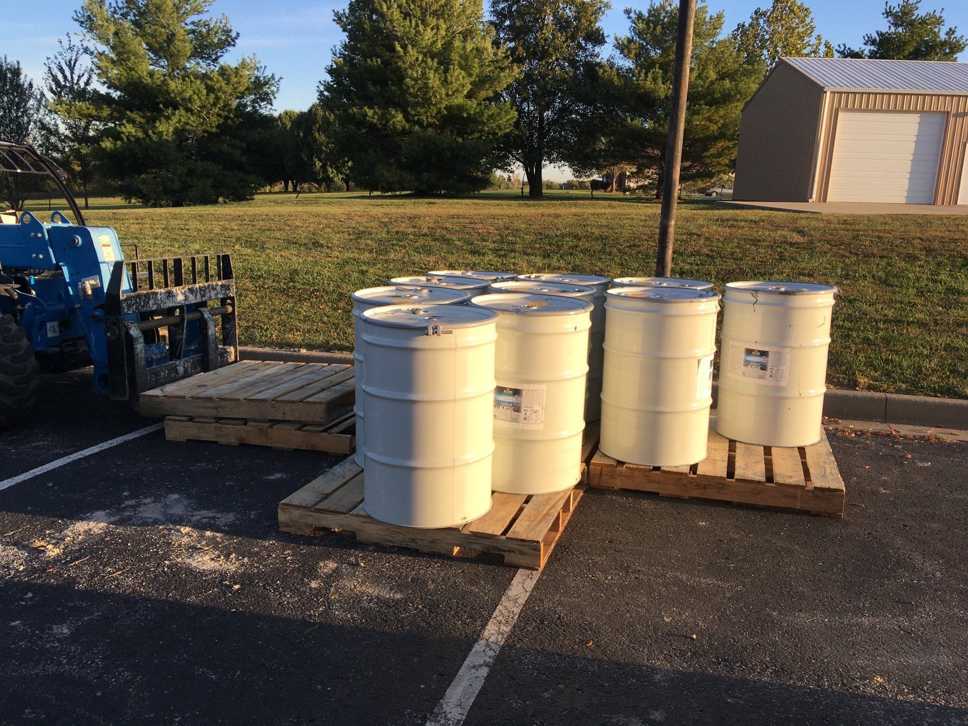 Several white barrels of roof coating are stacked on wooden pallets in a parking lot