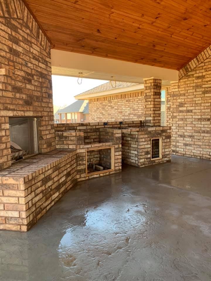 Covered brick patio with built-in fireplace and outdoor kitchen under a wood ceiling, wet concrete floor.