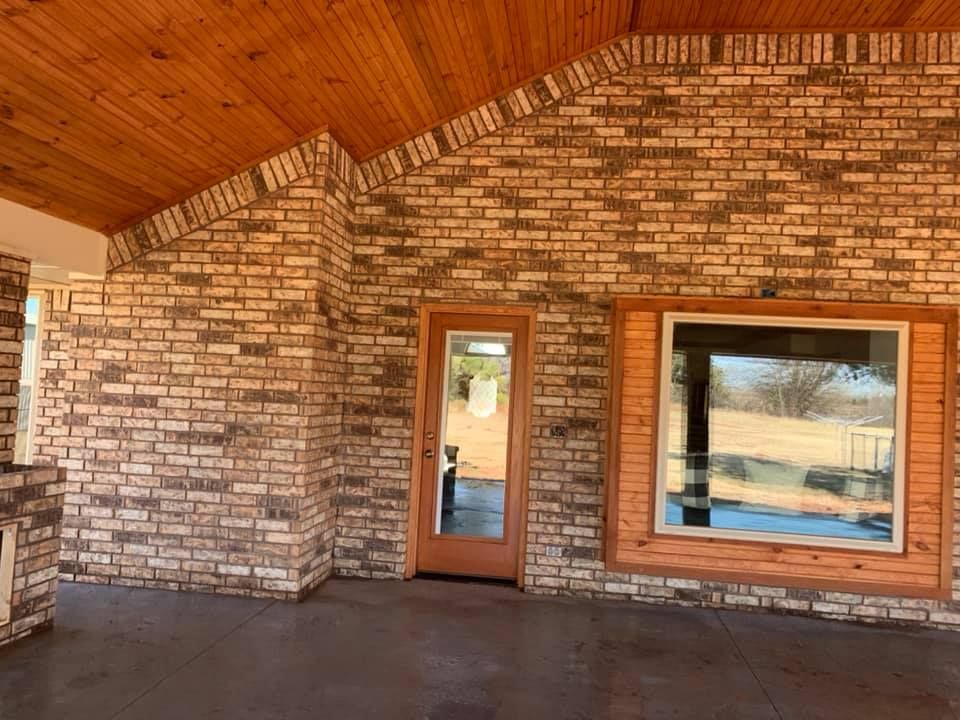 Covered brick patio with wood ceiling, glass door, and large window overlooking a pool area
