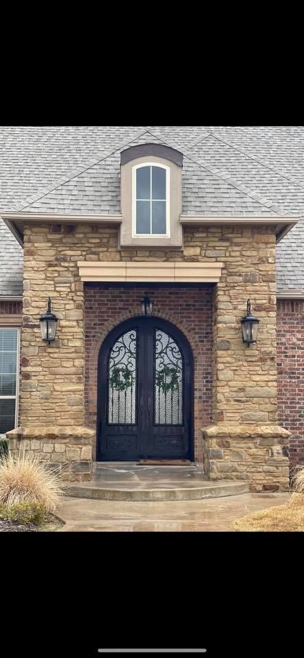 Stone house entryway with arched black double doors and lanterns beneath a dormer window