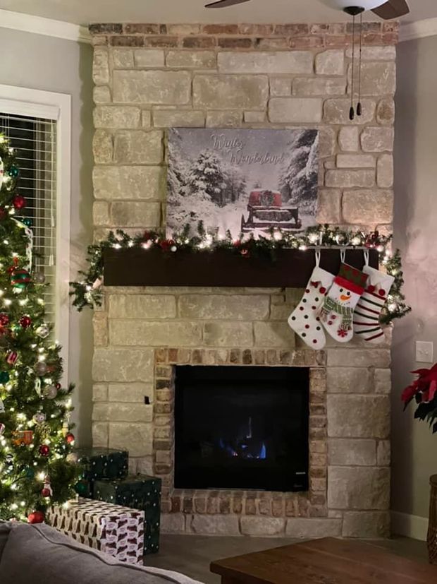 Stone fireplace decorated for Christmas with garland, stockings, and a holiday painting above the mantel.