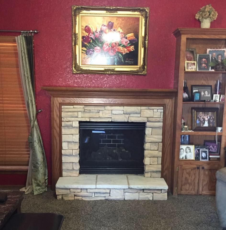 Living room fireplace with stone surround, red walls, floral painting, and wooden shelf with framed photos