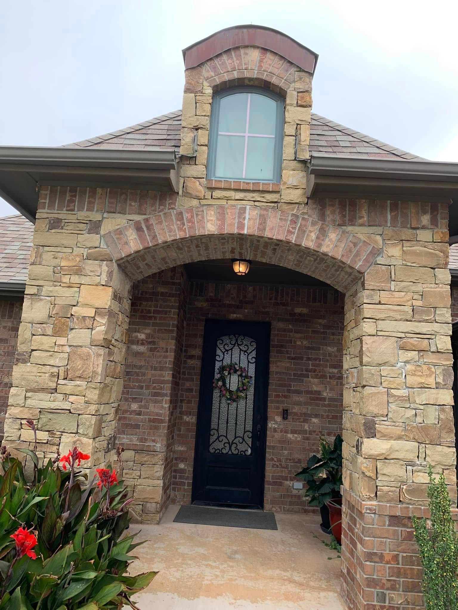 Stone house entry with arched porch, black front door, and flowers by the walkway