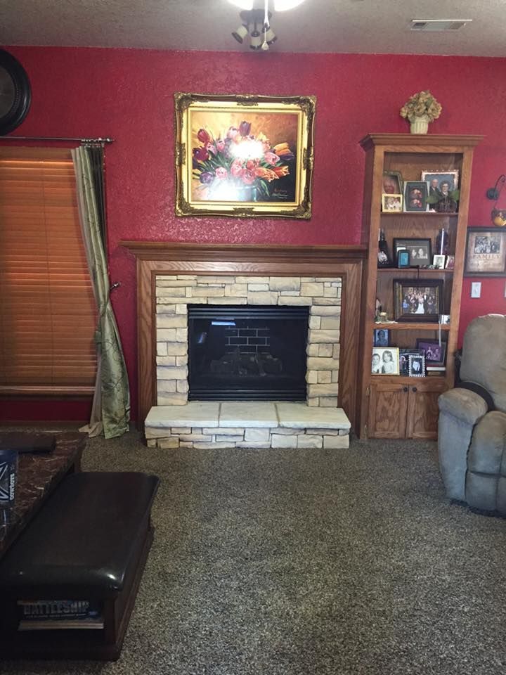 Living room with a brick fireplace, red walls, framed painting, shelves, and carpeted floor.