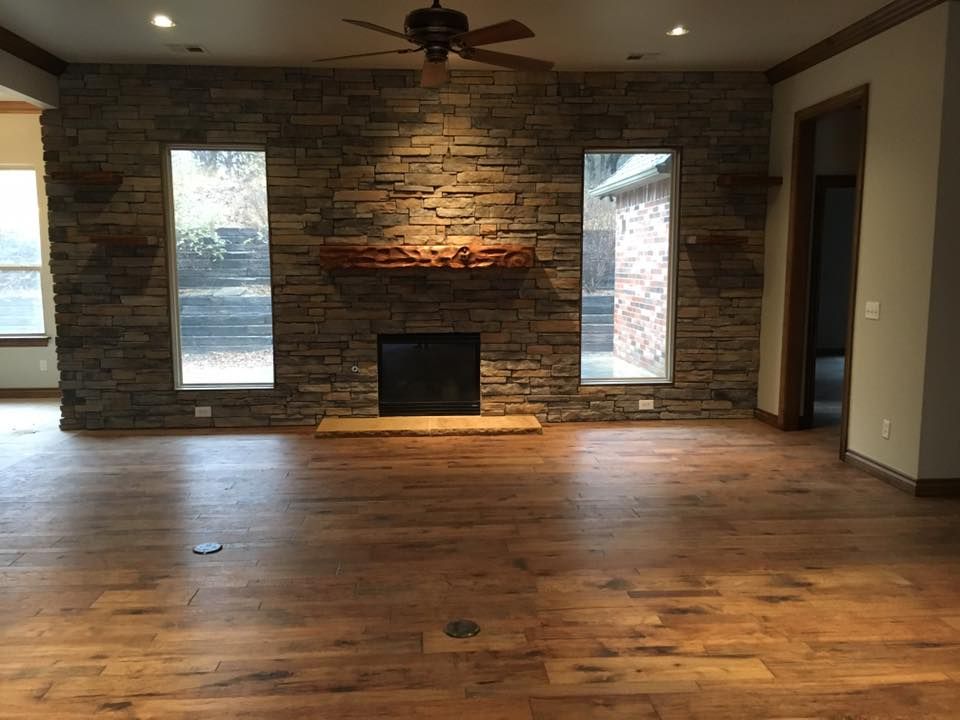 Empty living room with hardwood floor, stone fireplace wall, ceiling fan, and tall windows.