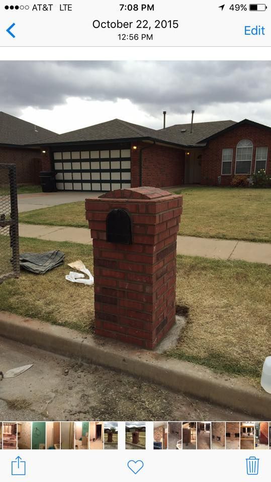Brick mailbox by a driveway in front of a house with a garage, on a grassy suburban lawn.