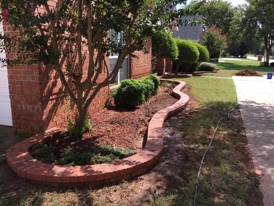 Curved red-brick garden border beside a brick house, with shrubs and a driveway next to it