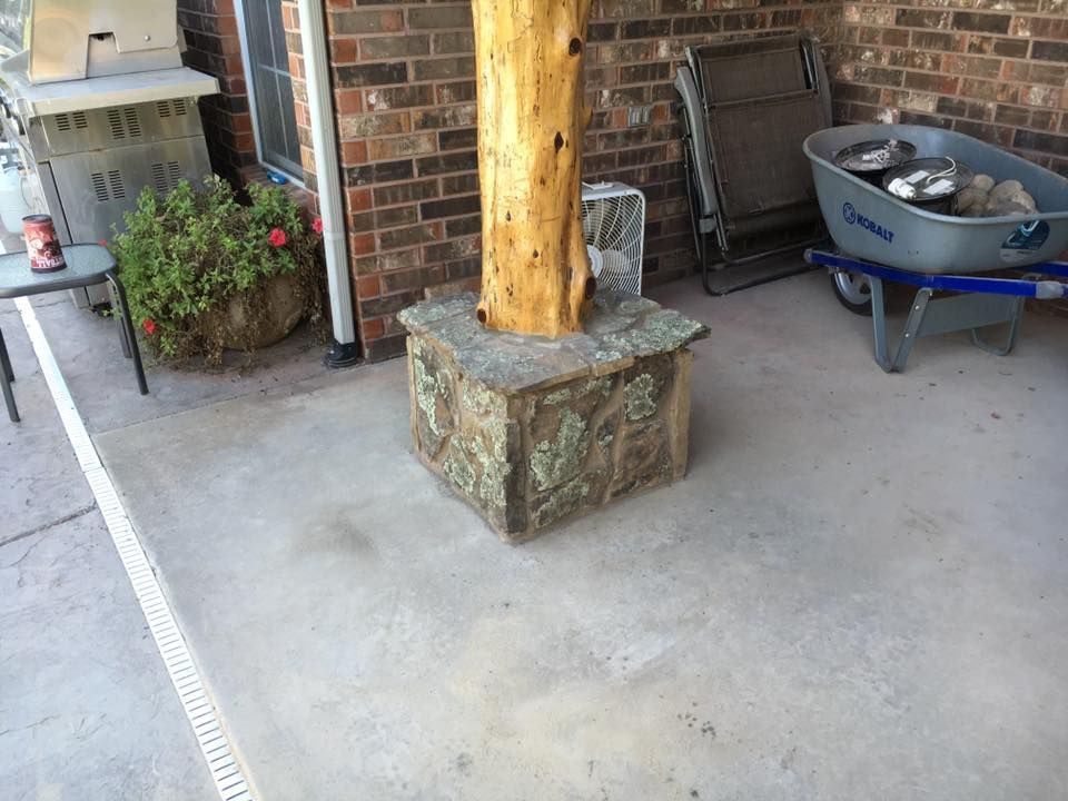 Porch with stone column base, concrete floor, potted plants, and a blue wheelbarrow of rocks.
