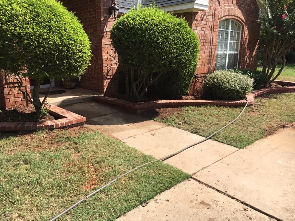 Brick house entrance with concrete walkway, trimmed bushes, and green lawn in front yard
