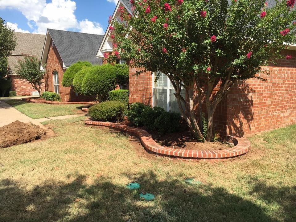 Brick house yard with flowering tree, trimmed shrubs, and a small lawn in sunlight