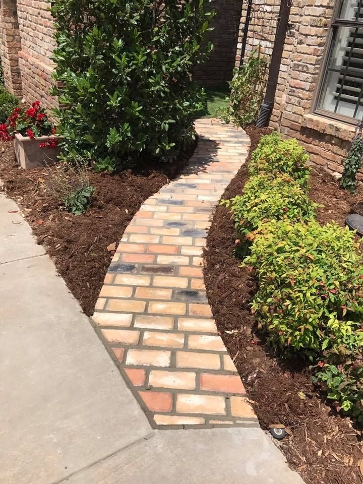 Curved brick garden path beside a concrete walkway, bordered by mulch and shrubs near a house wall.