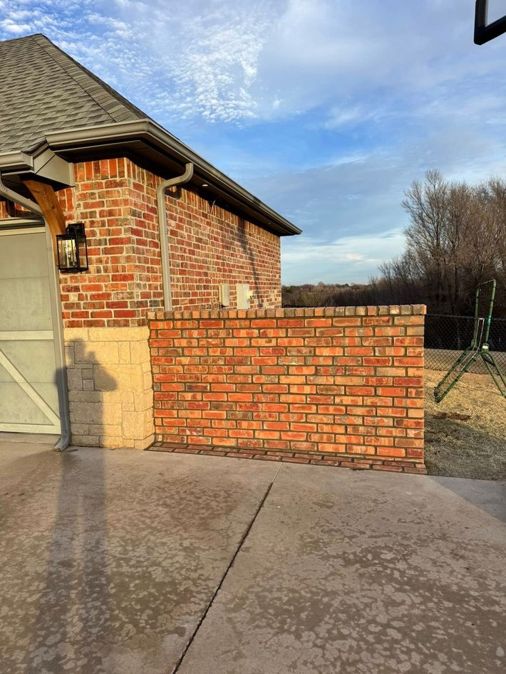 Brick wall beside a brick house and driveway under a blue sky, with a fenced yard in the background
