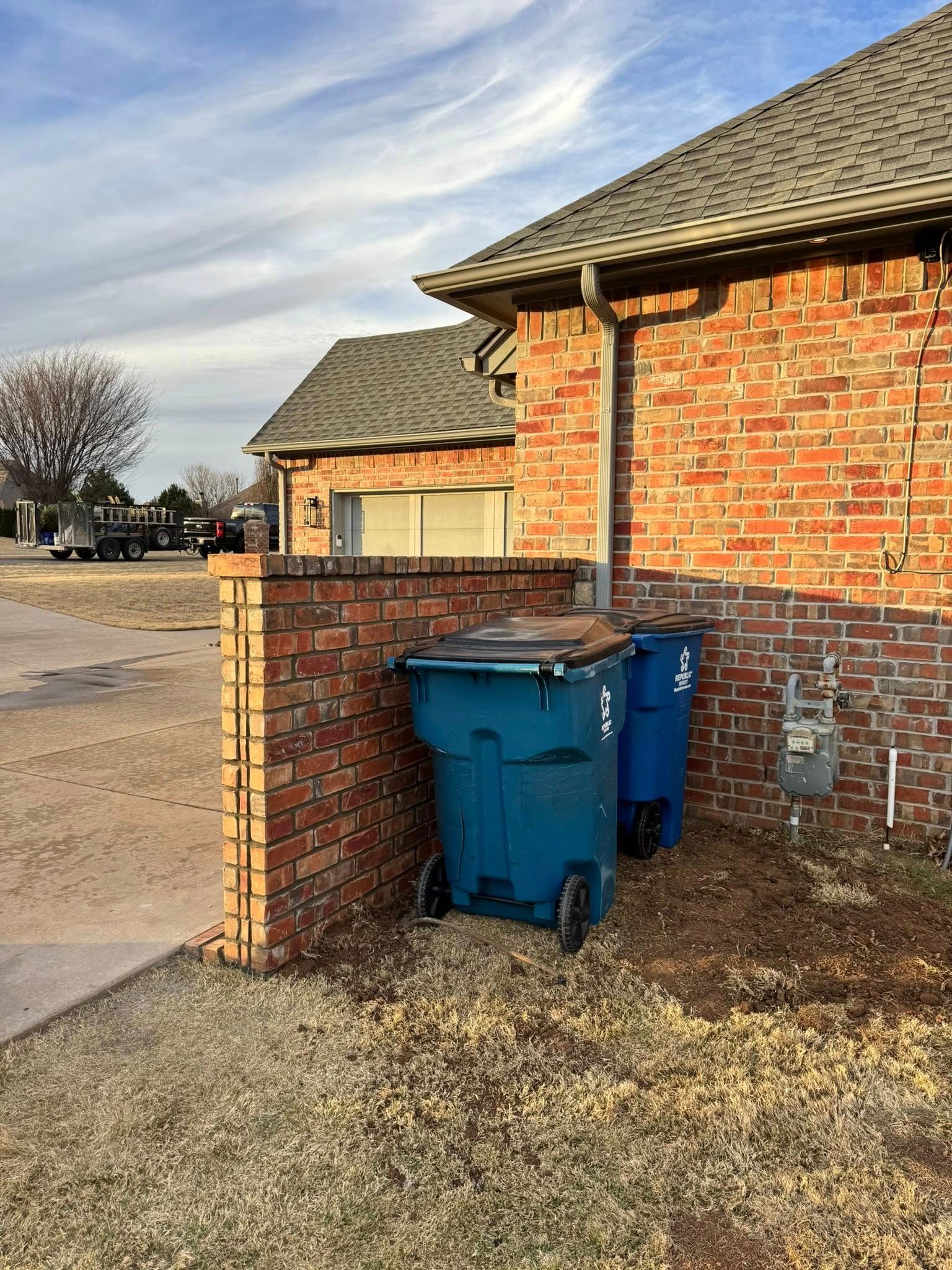 Blue trash bins beside a brick house on a gravel driveway under a cloudy sky