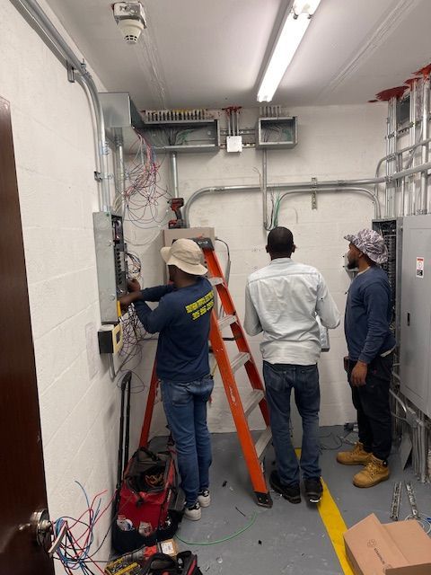 Three electricians working on wiring in a utility room. One on a ladder, two others observing.