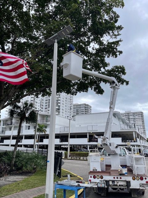 Man in bucket truck working on a tall light pole with solar panel in front of buildings.
