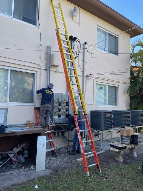 Two workers on ladders install electrical components on a building exterior.