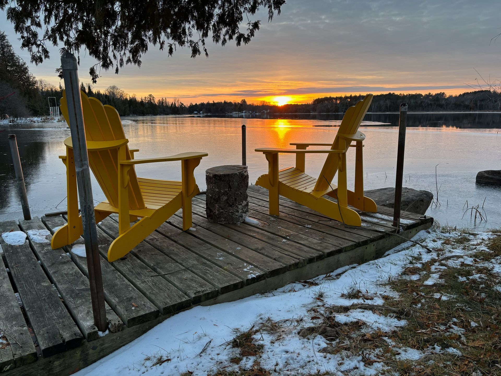 Two yellow adirondack chairs are sitting on a dock overlooking a lake at sunset.