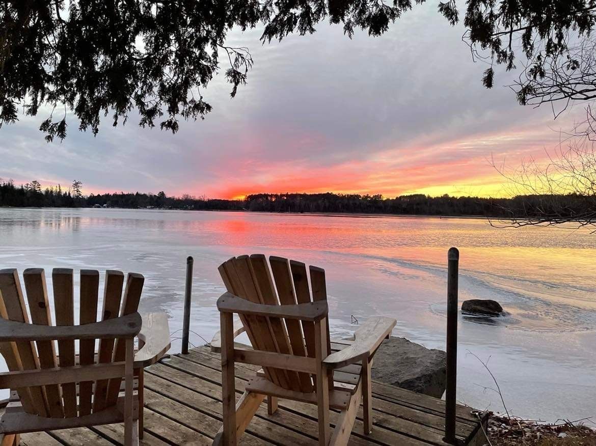 Two wooden chairs are sitting on a dock overlooking a lake at sunset.