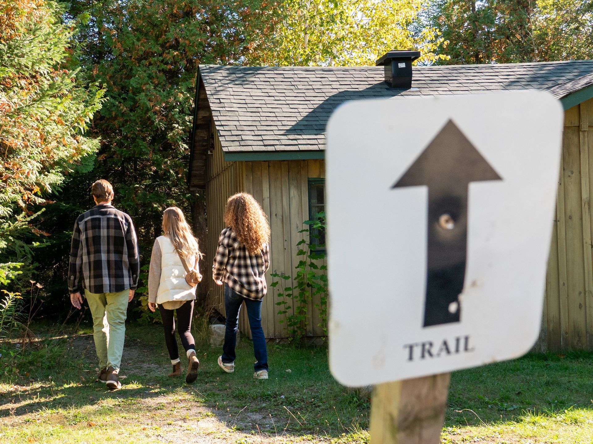 Three people walk toward a trail sign with an arrow pointing forward, next to a cabin.