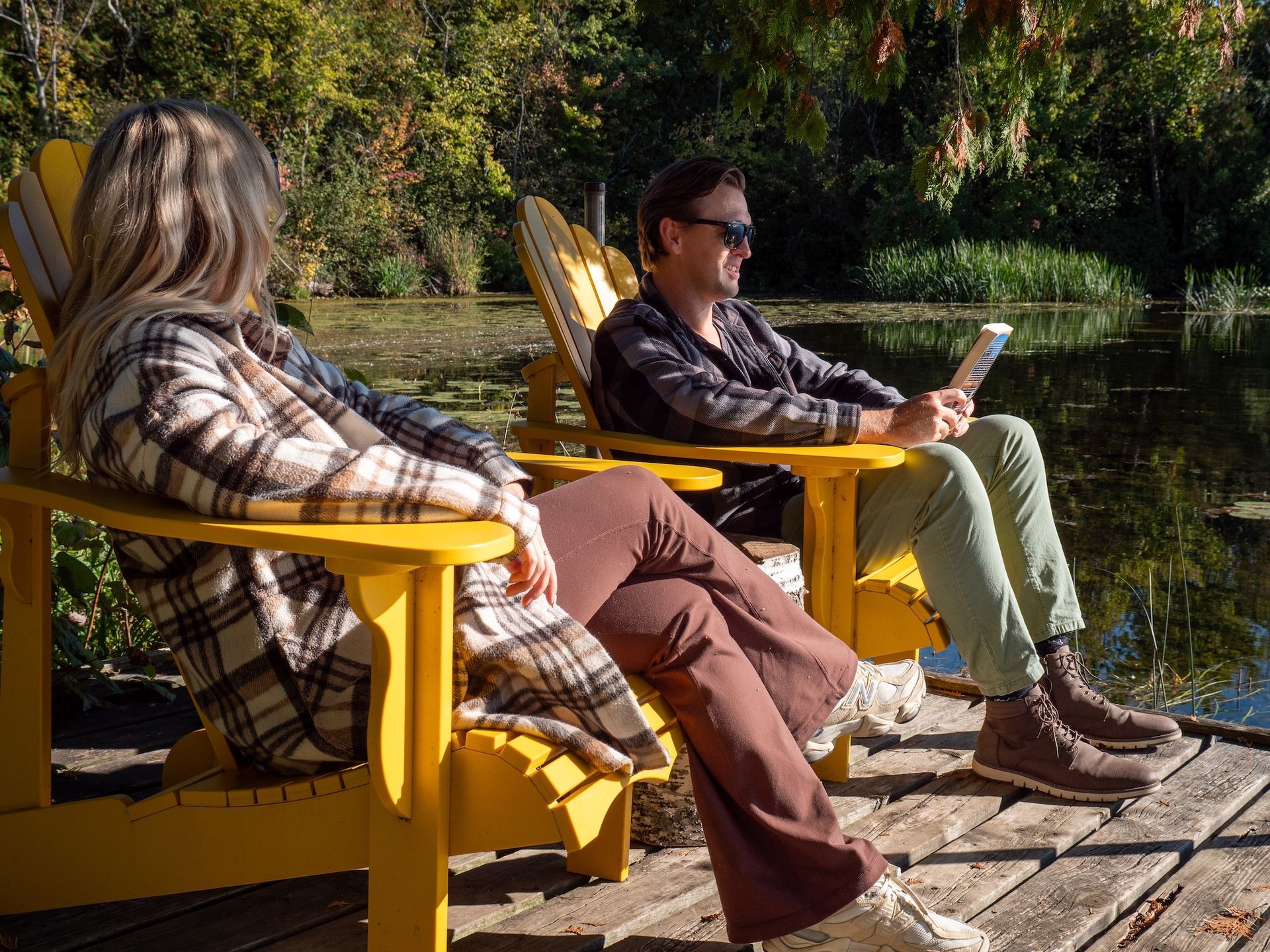 Two people relax in yellow Adirondack chairs by a pond. One reads a book, the other looks out. Sunny day.