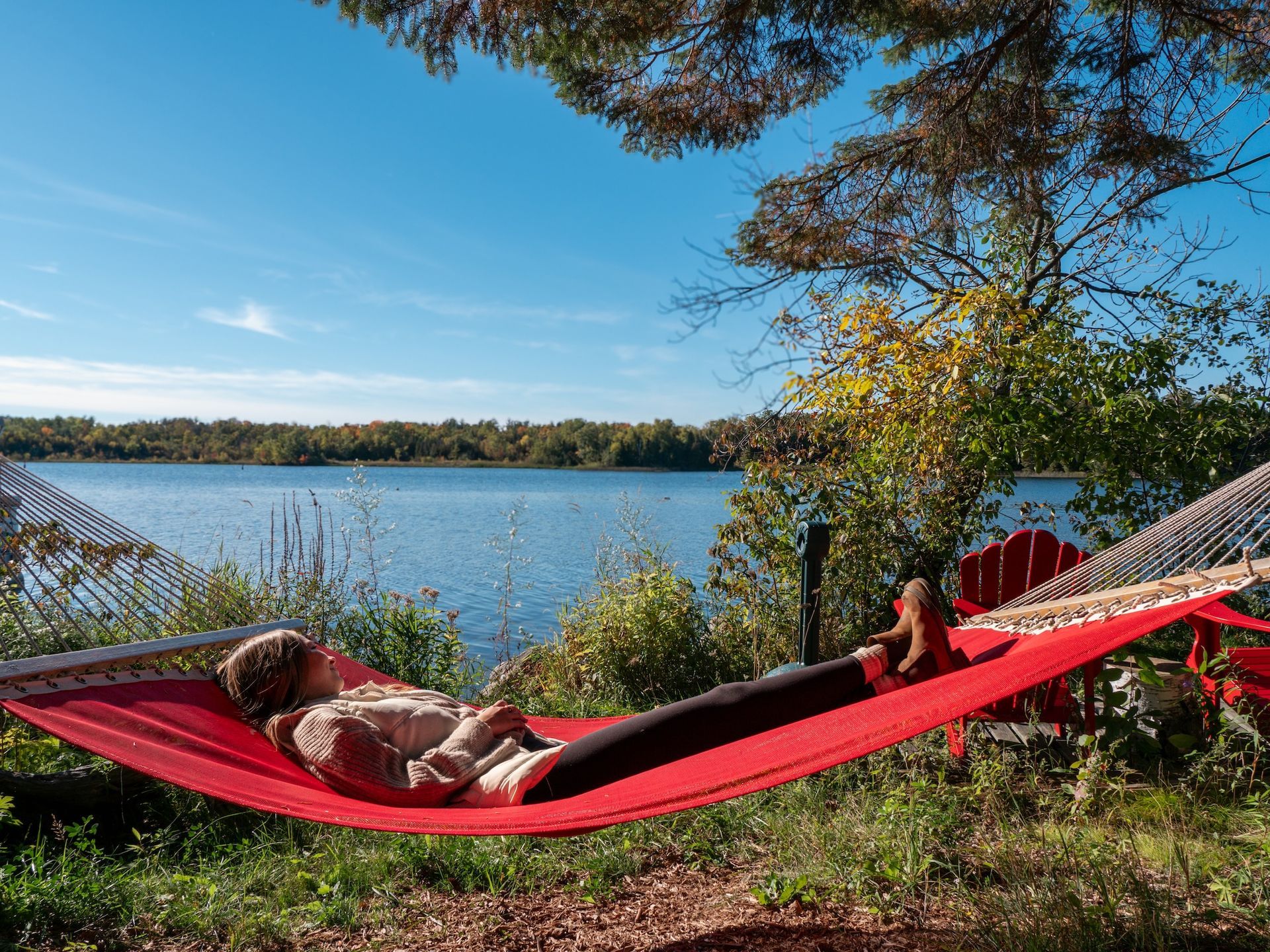 Person resting in a red hammock by a lake on a sunny day.