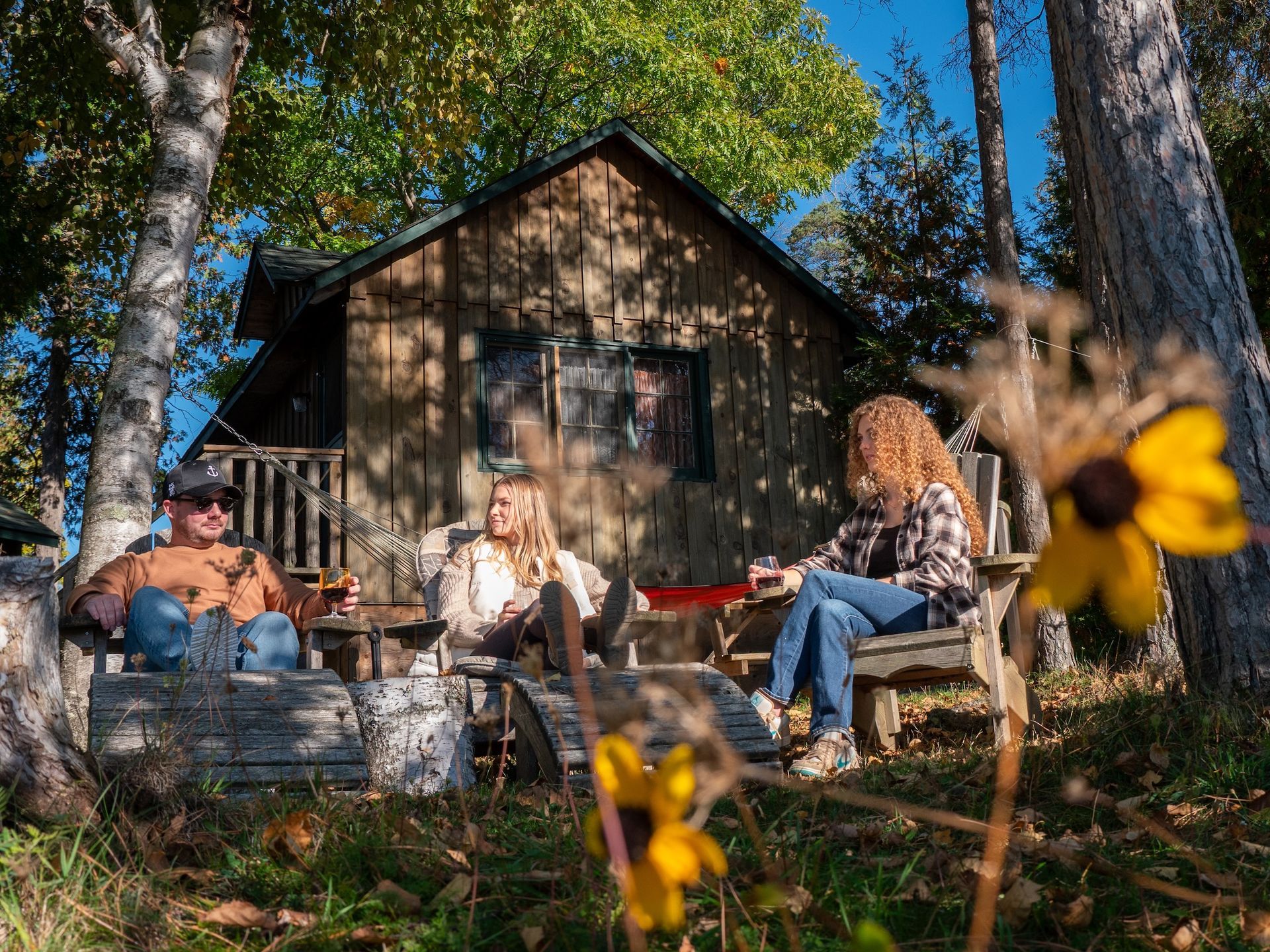 Three people sit in chairs outside a wooden cabin; flowers in the foreground.