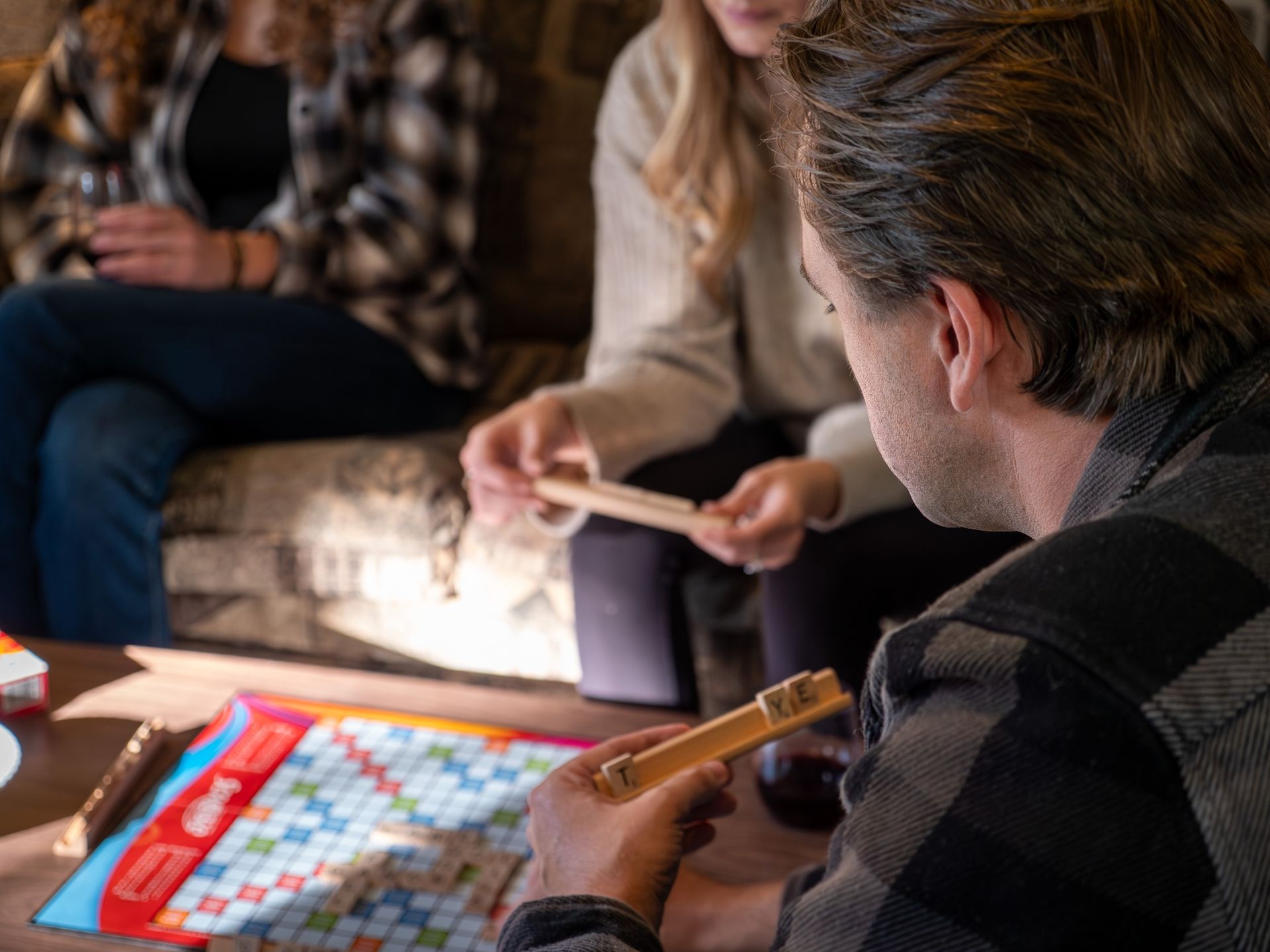 People playing Scrabble on a coffee table. One man holds tiles; two others look on.