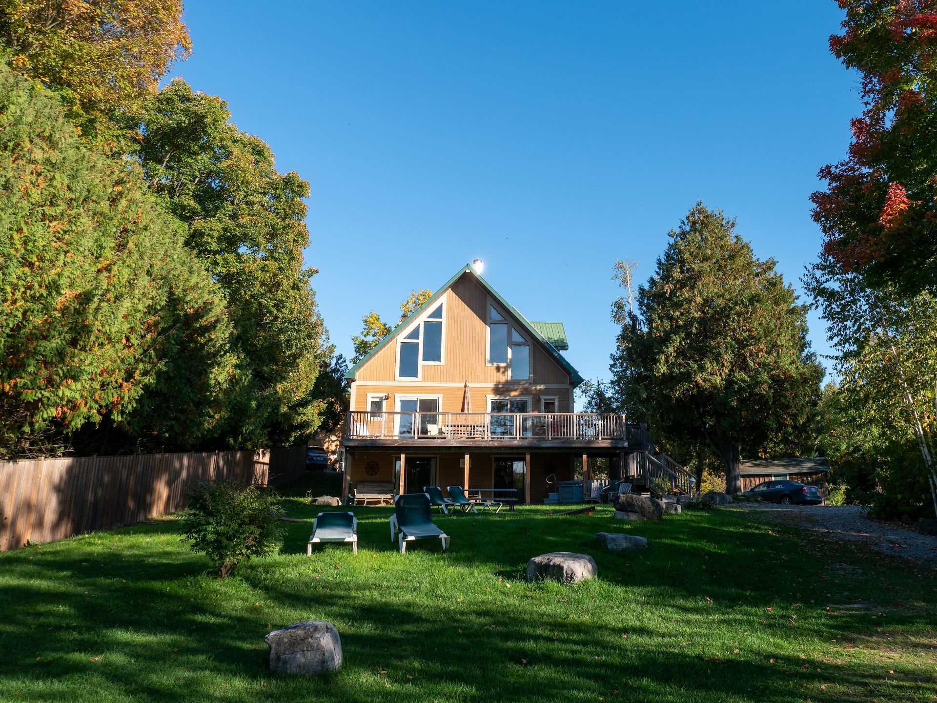House with deck on a grassy lawn, flanked by trees under a blue sky; lounge chairs sit on the grass.