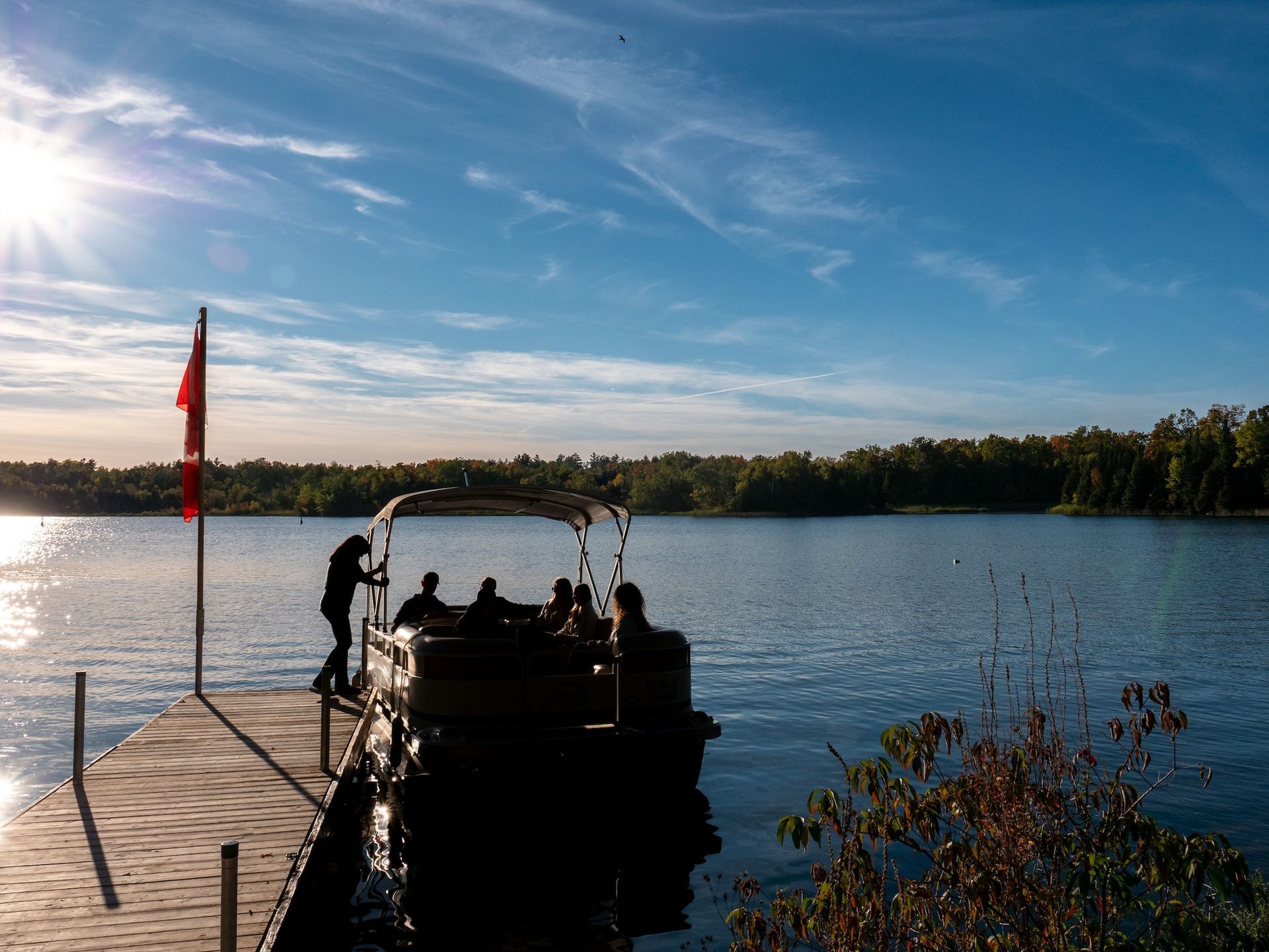 Sunlit lake scene with pontoon boat on a dock; figures boarding; trees and blue sky.
