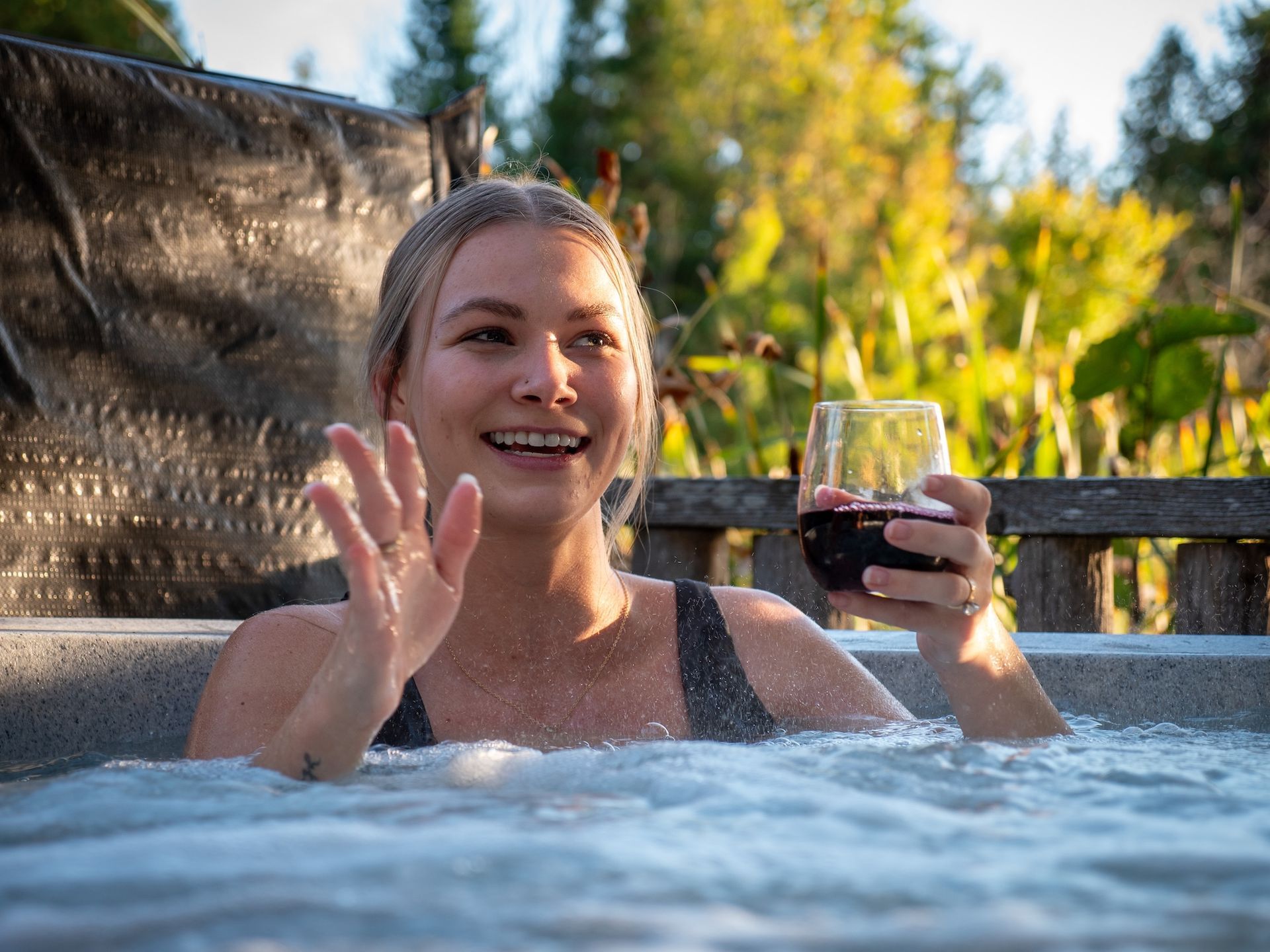 Woman in a hot tub, waving and holding a glass of red wine, outdoors in sunlight.