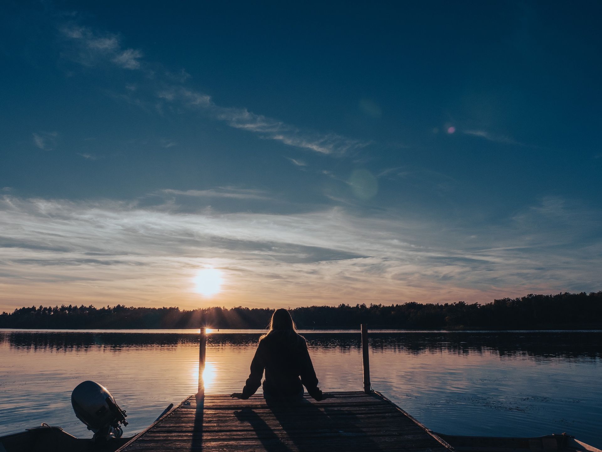 Woman silhouetted on a wooden dock, facing a sunset over a calm lake.