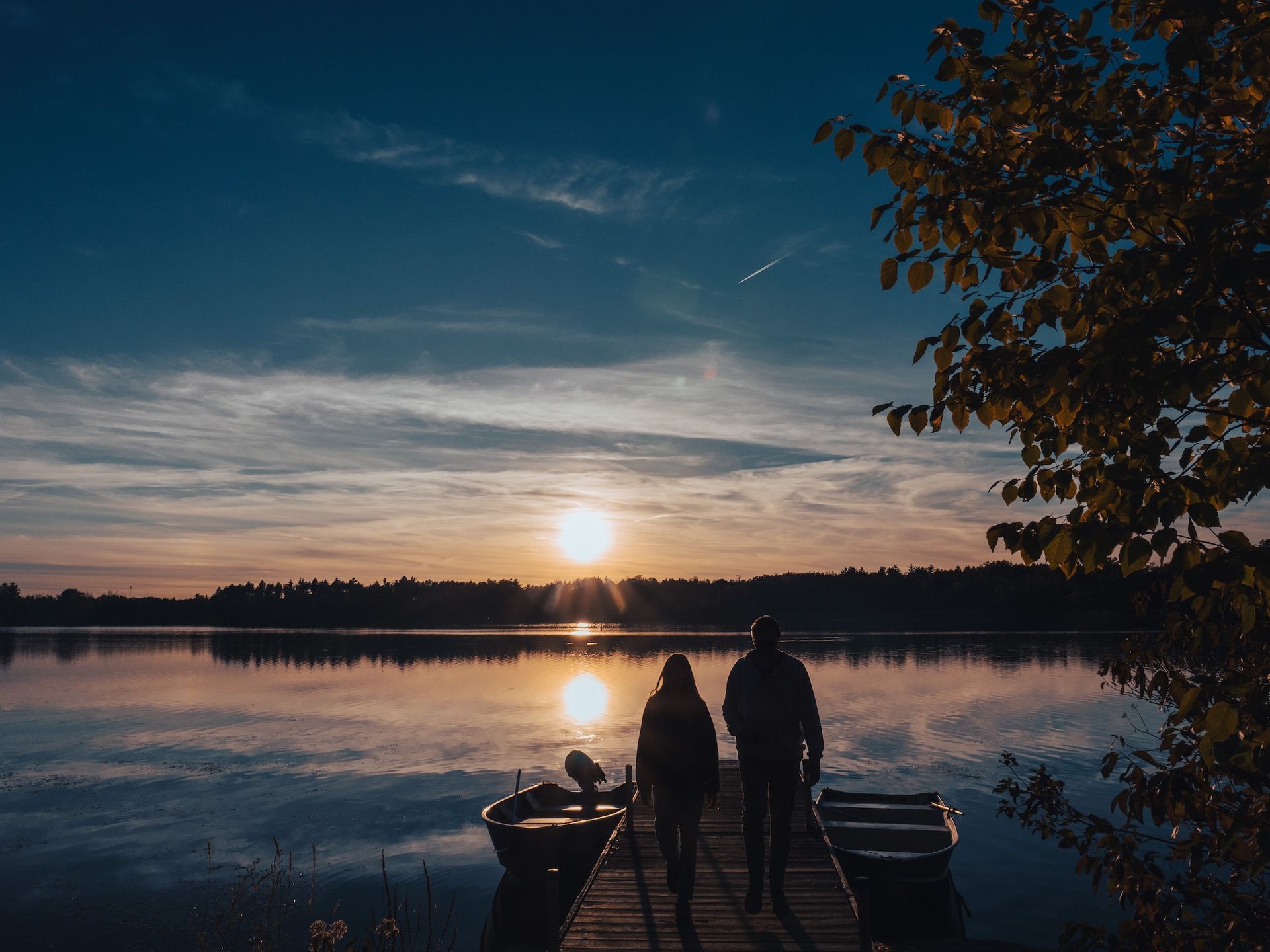 Couple walking on a dock at sunset, overlooking calm lake. Boats are moored; trees line the opposite shore.