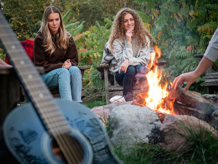 Two people sitting by a campfire with a guitar in the foreground. They are outdoors, with foliage.