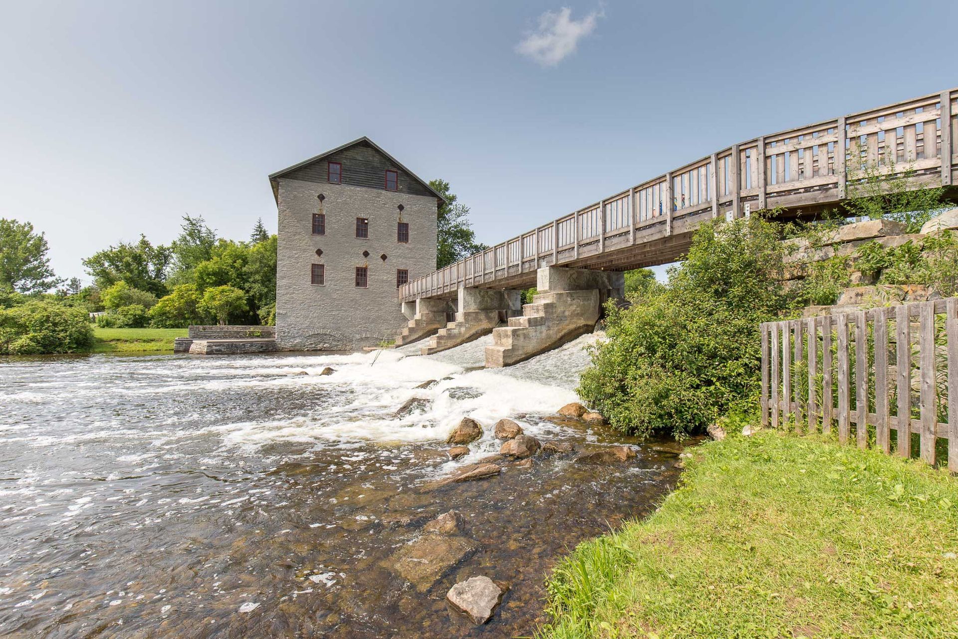 Stone mill building next to a flowing river with a wooden bridge and grassy banks under a blue sky.