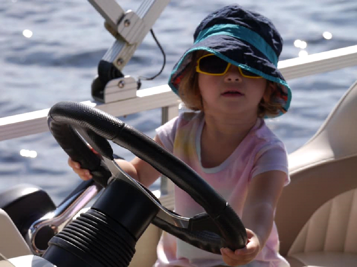 A little girl wearing a hat and sunglasses is driving a boat