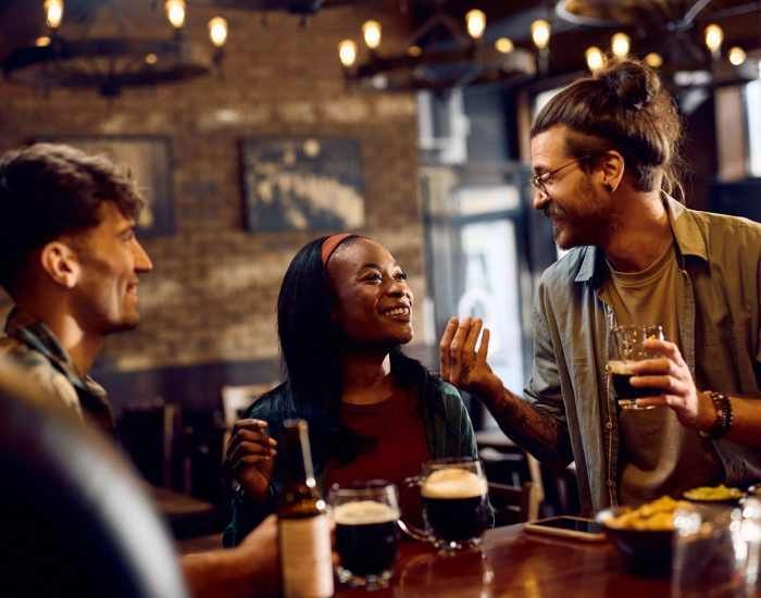 Three people at a bar, laughing and talking while holding drinks. Dark setting with overhead lights.