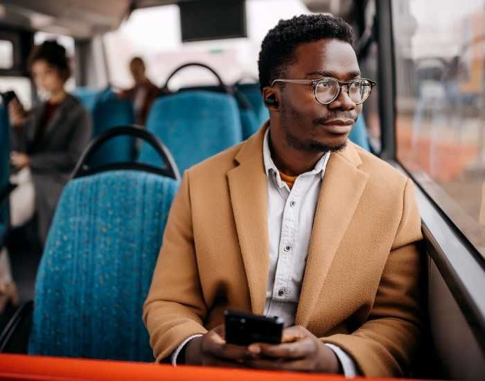 Man in glasses and tan coat looks out window on a bus, holding phone.