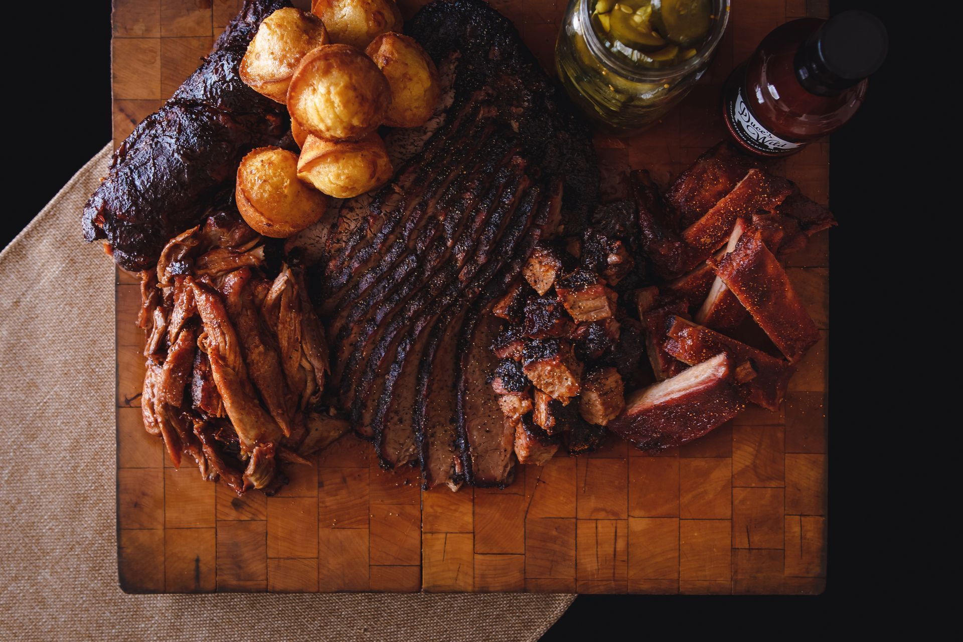 a platter of meats including pulled pork, brisket, and ribs with cornbread and pickles