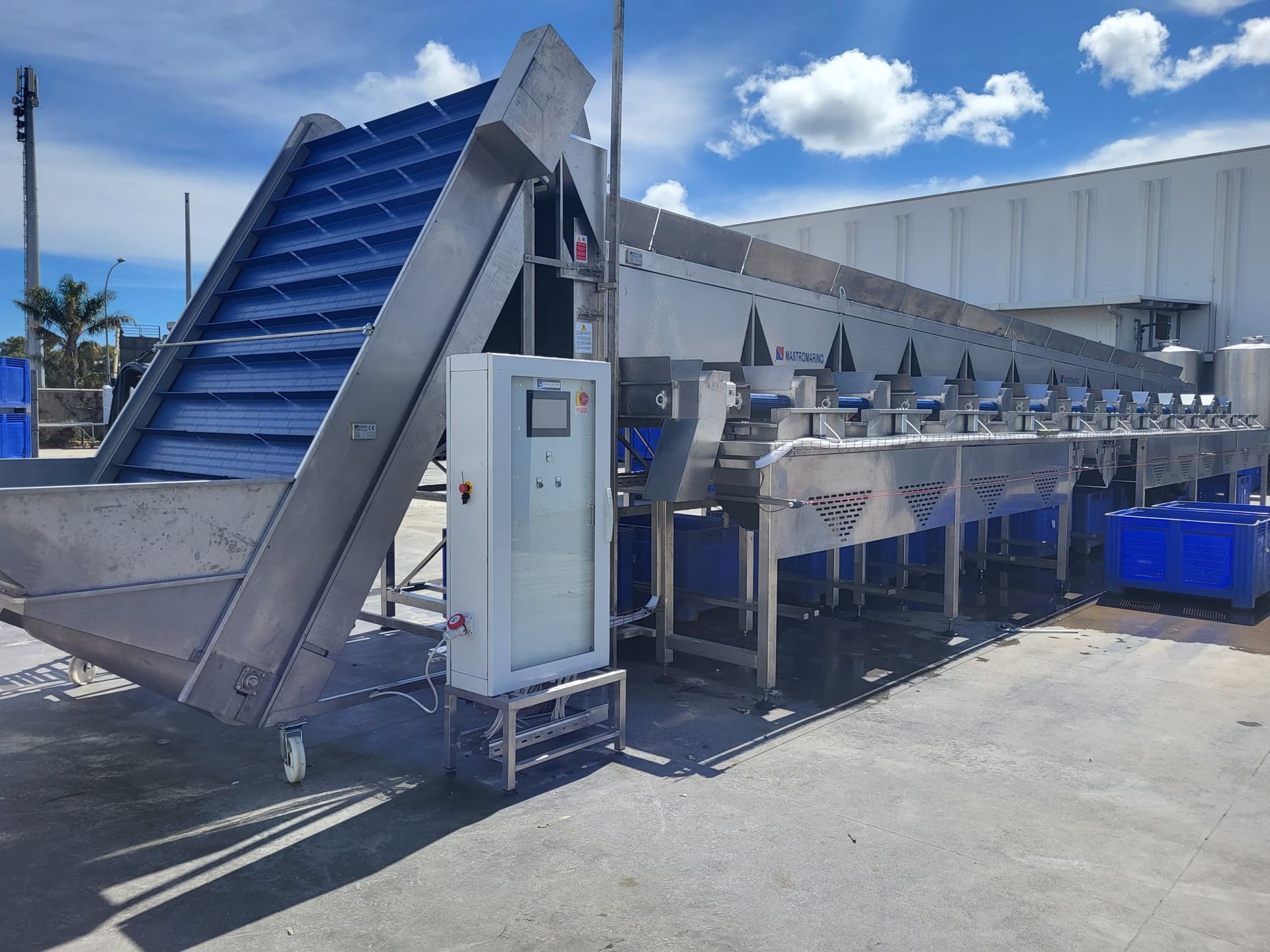 Macchina per la lavorazione degli alimenti in acciaio inossidabile con nastro trasportatore blu, all'aperto vicino a un magazzino in una giornata di sole.