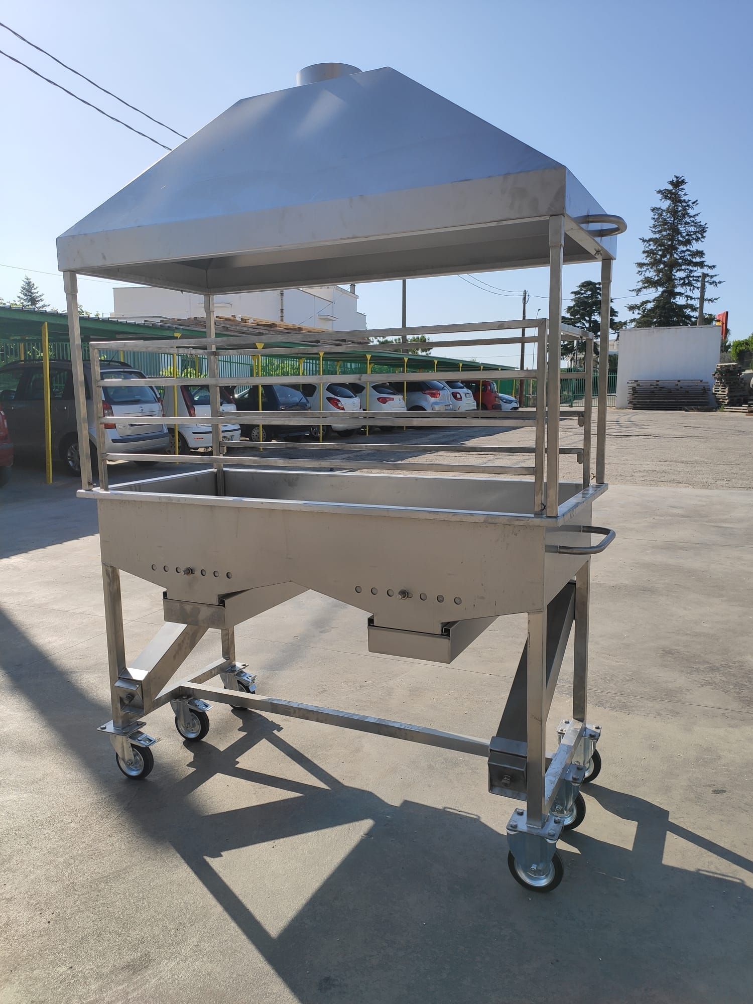 Grande barbecue da esterno in acciaio inox su ruote, con tettoia. All'aperto con parcheggio sullo sfondo.