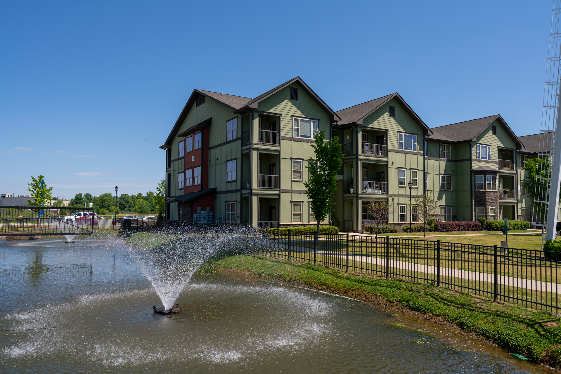 A green three-story apartment building with a water fountain in a pond out front under a clear blue sky at Riverwalk Apartments.