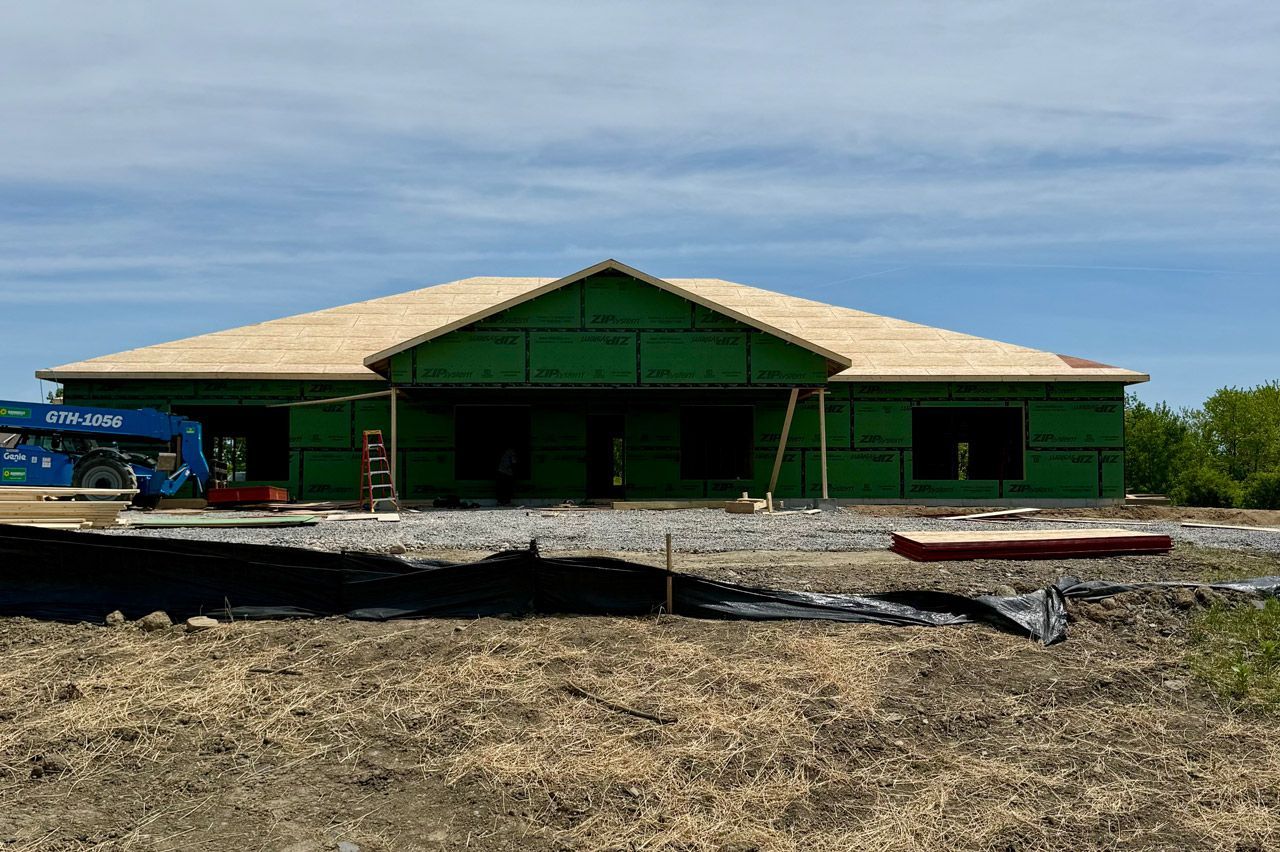Green house under construction on a sandy lot, with unfinished roof and open front under a blue sky.